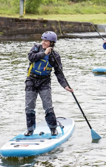 Two people stand-up paddleboarding on a calm canal, both wearing helmets and buoyancy aids. Two people stand-up paddleboarding on a calm canal, both wearing helmets and buoyancy aids.